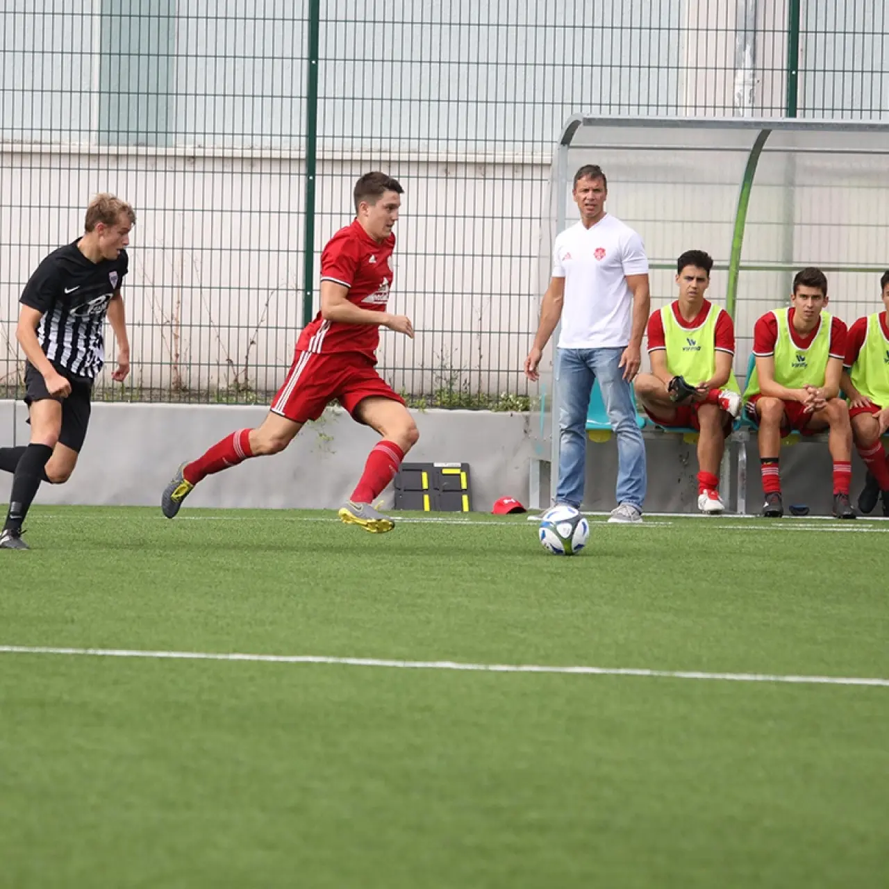 Rotgekleideter Spieler treibt den Ball über den Kunstrasen, verfolgt von einem Gegner in Schwarz-Weiß, im Hintergrund die Ersatzbank und ein Betreuer.