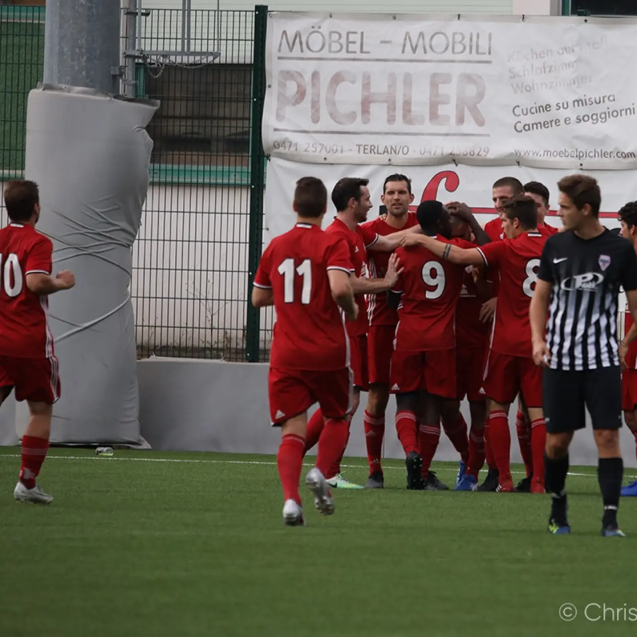 Rot gekleidete Spieler des AFC Terlan jubeln in einer Traube auf dem Kunstrasen, im Hintergrund Bande des Sponsors Möbel Pichler.