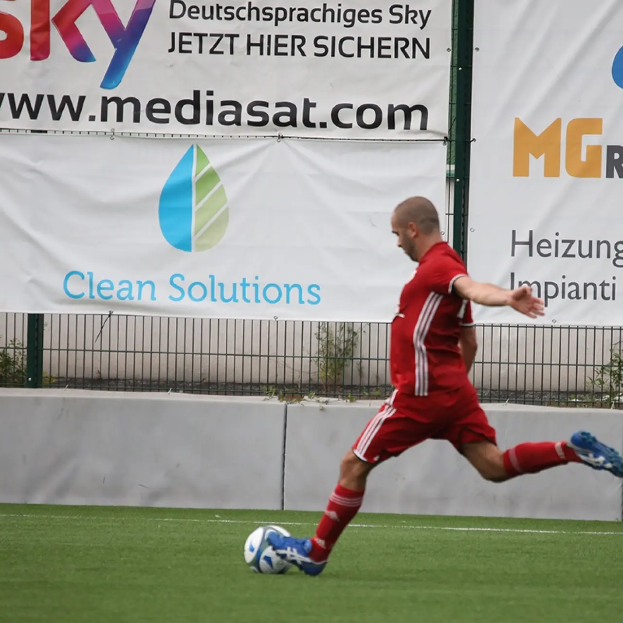 Ein Spieler in roter Trikotkombination schießt auf dem Kunstrasen den Ball, im Hintergrund Werbebanden von MG Rainer und Clean Solutions.