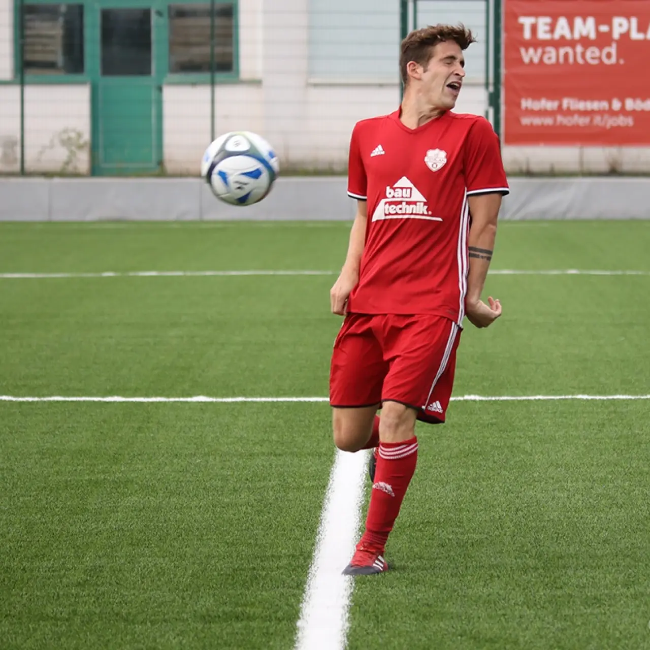 Spieler in roten Trikot und Stutzen kontrolliert den Ball mit dem Knie auf dem Kunstrasen, im Hintergrund Bandenwerbung von Hofer Fliesen & Böden.