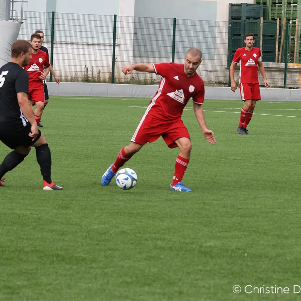 Spieler in roten Trikots führt den Ball am Mittelkreis, während ein Gegenspieler in Schwarz zu ihm aufschließt; im Hintergrund weitere Spieler und ein Schiedsrichter in Neongelb.