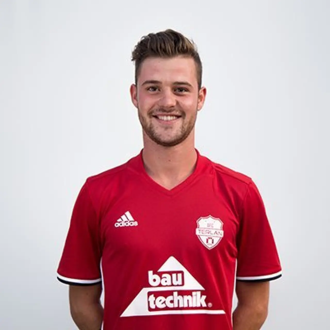 Porträt eines lächelnden jungen Fußballspielers in rotem AFC-Terlan-Trikot mit Bautechnik-Sponsorenlogo vor weißem Hintergrund.