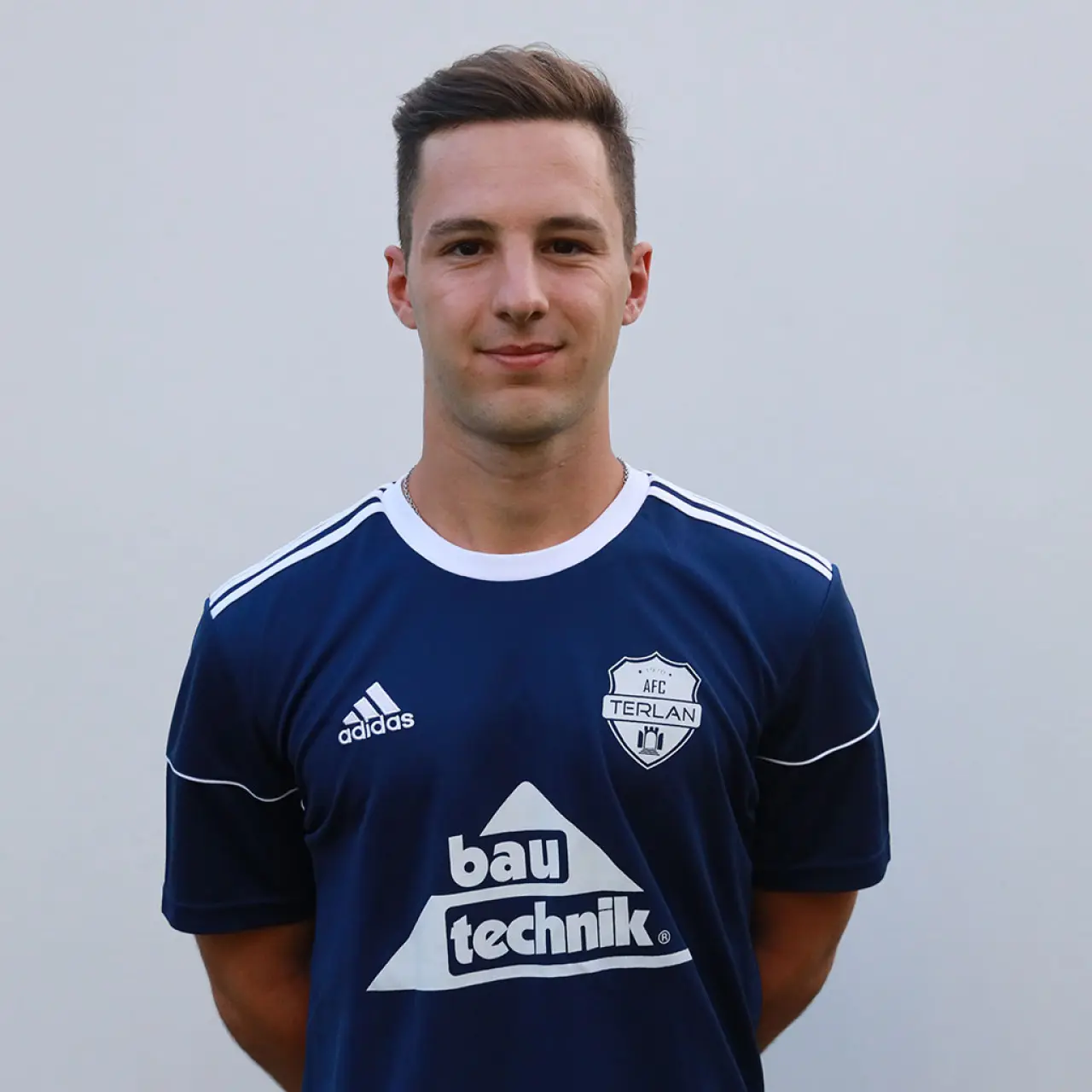 Porträtfoto eines jungen Mannes im dunkelblauen AFC-Terlan-Trikot mit Vereinswappen und bautechnik-Sponsorenlogo vor einer hellen Wand.
