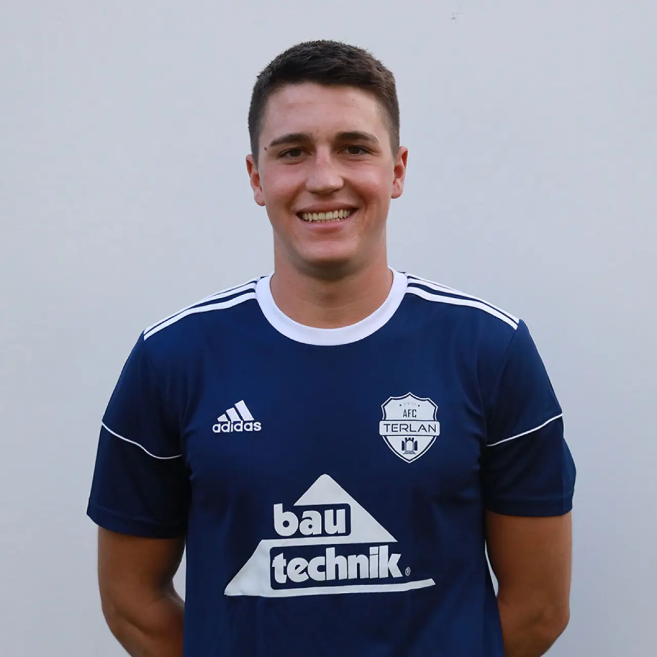 Porträt eines lächelnden jungen Spielers im dunkelblauen AFC-Terlan-Trikot mit bautechnik-Sponsorenaufdruck vor einer hellgrauen Wand.