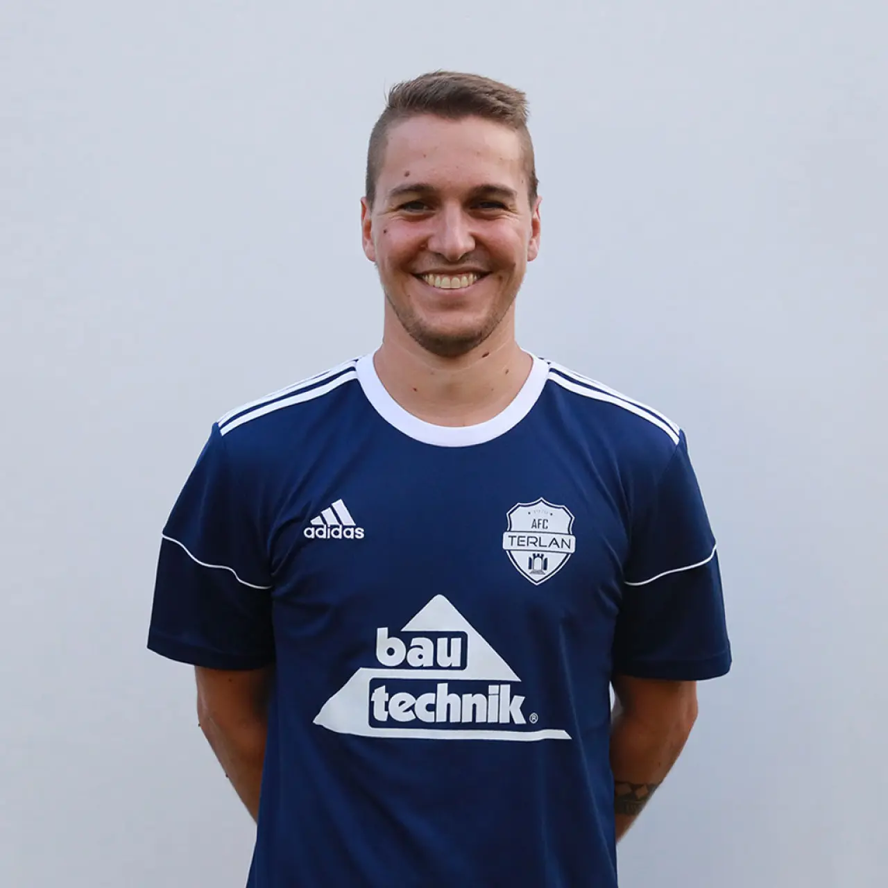 Porträt eines lächelnden Spielers im dunkelblauen AFC-Terlan-Trikot mit bautechnik-Sponsorenaufdruck vor grauem Hintergrund.