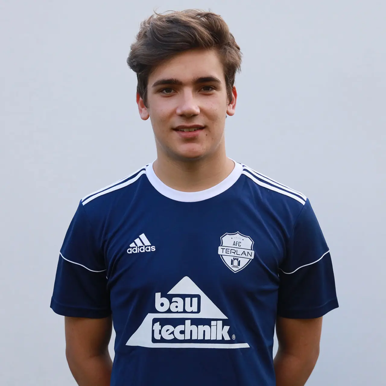 Porträt eines jungen Fußballspielers im dunkelblauen AFC-Terlan-Trikot mit Bautechnik-Sponsor vor neutral grauem Hintergrund.