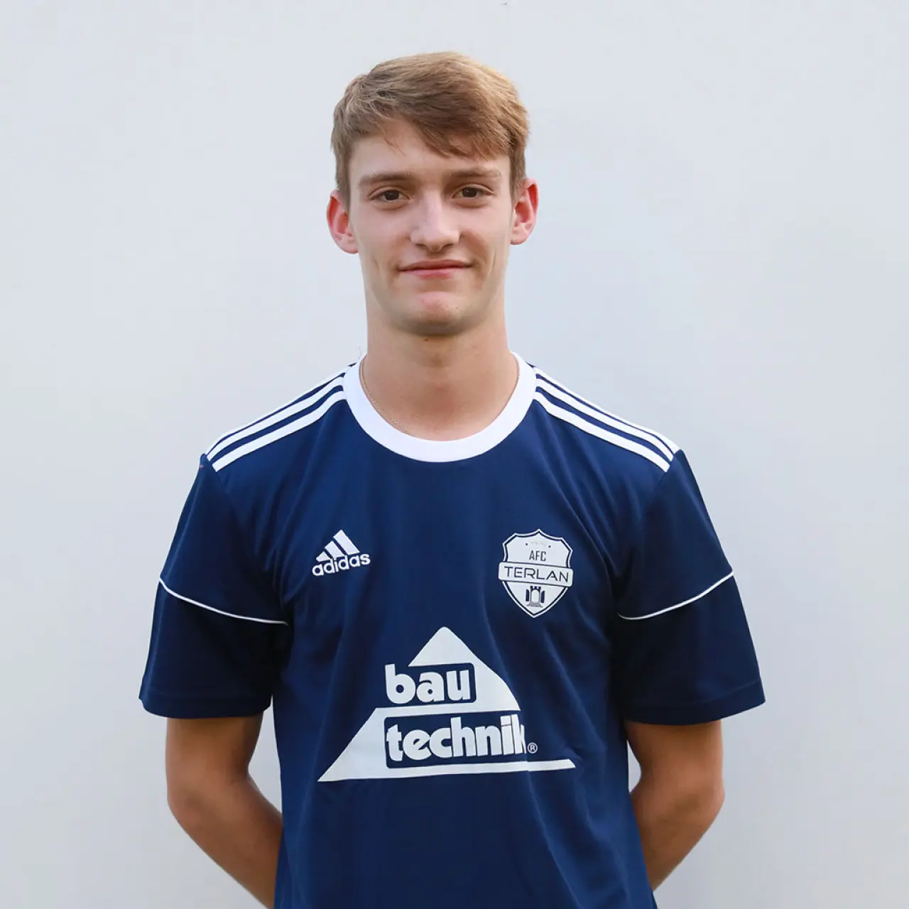 Porträtfoto eines jungen Fußballspielers im dunkelblauen AFC-Terlan-Trikot mit Bautechnik-Sponsorlogo vor einer hellgrauen Wand.