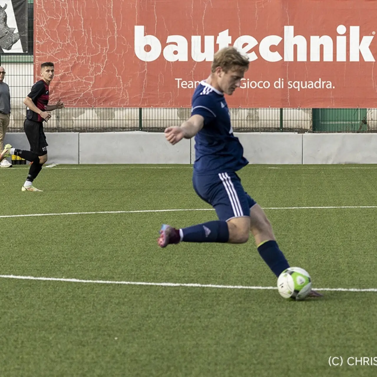 Spieler in dunkelblauem Trikot schießt beim Spiel des AFC Terlan gegen Partschins den Ball auf dem Kunstrasen, im Hintergrund Bande mit Bautechnik-Werbung.