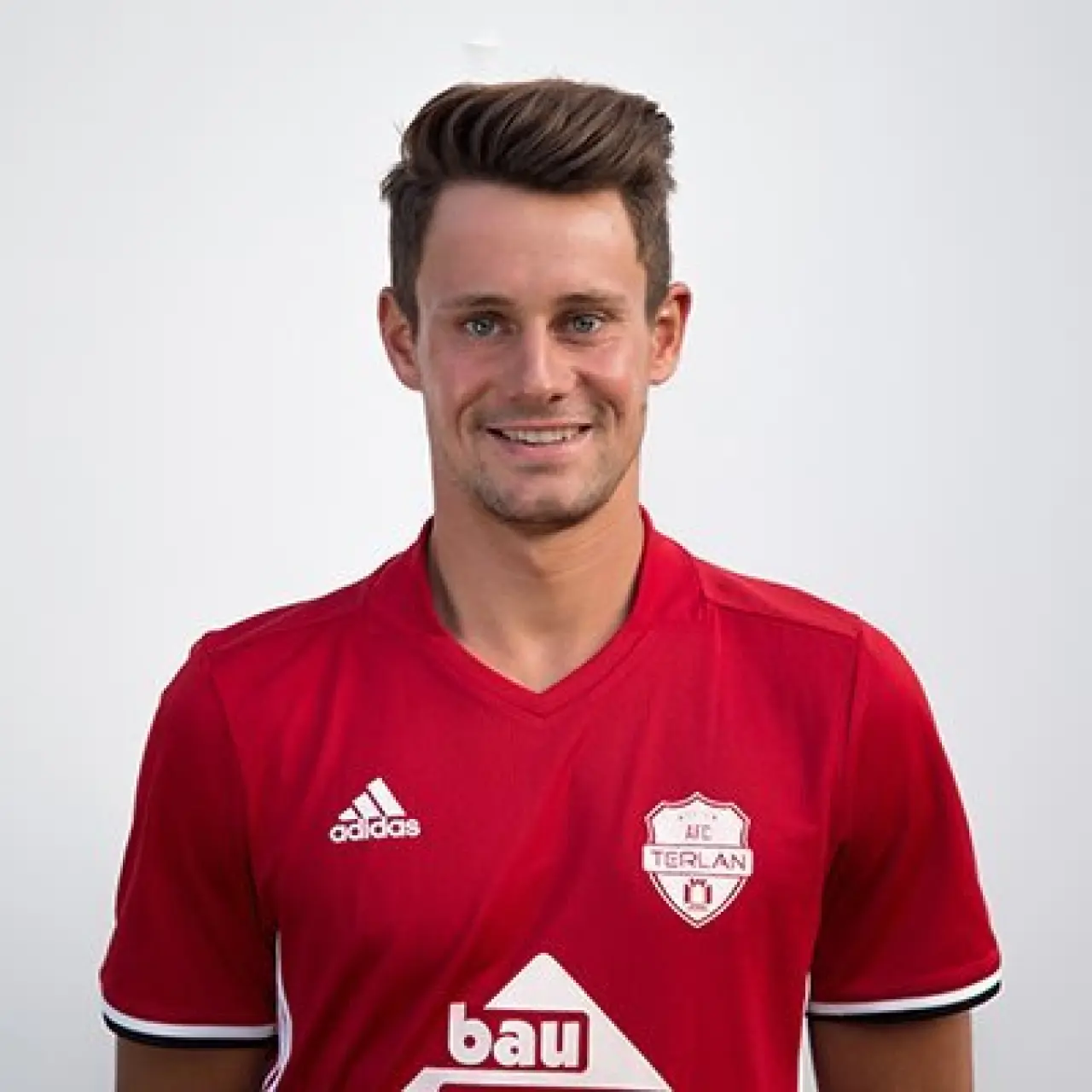 Porträt eines lächelnden Spielers des AFC Terlan in rotem Trikot mit Vereinswappen und Adidas-Logo vor hellem Hintergrund.