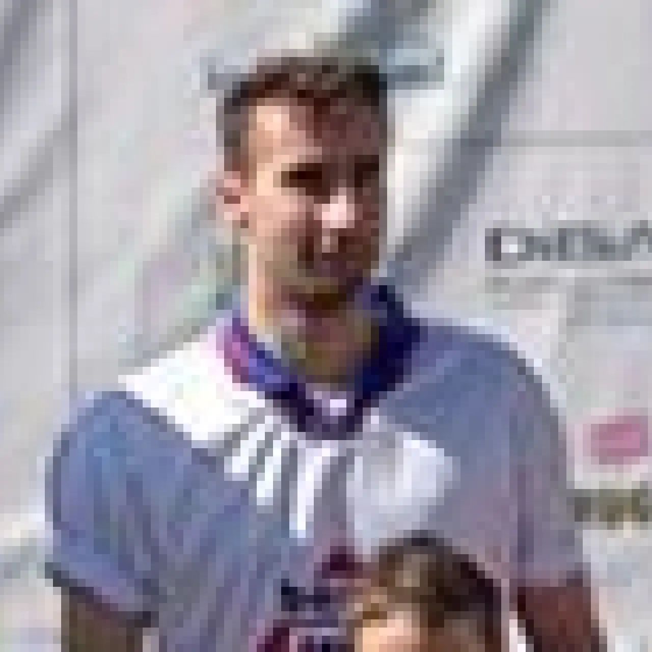 Ein Spieler in weißem Trikot mit violetten Akzenten blickt in die Kamera, im Hintergrund eine Sportanlage mit Werbebande.