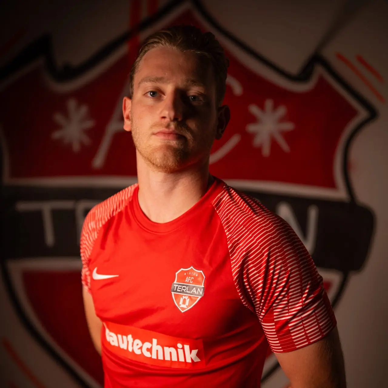 Spieler des AFC Terlan im roten Trikot mit Vereinswappen und Sponsorlogo vor einer großen Wappen-Wand, ernst in die Kamera blickend.