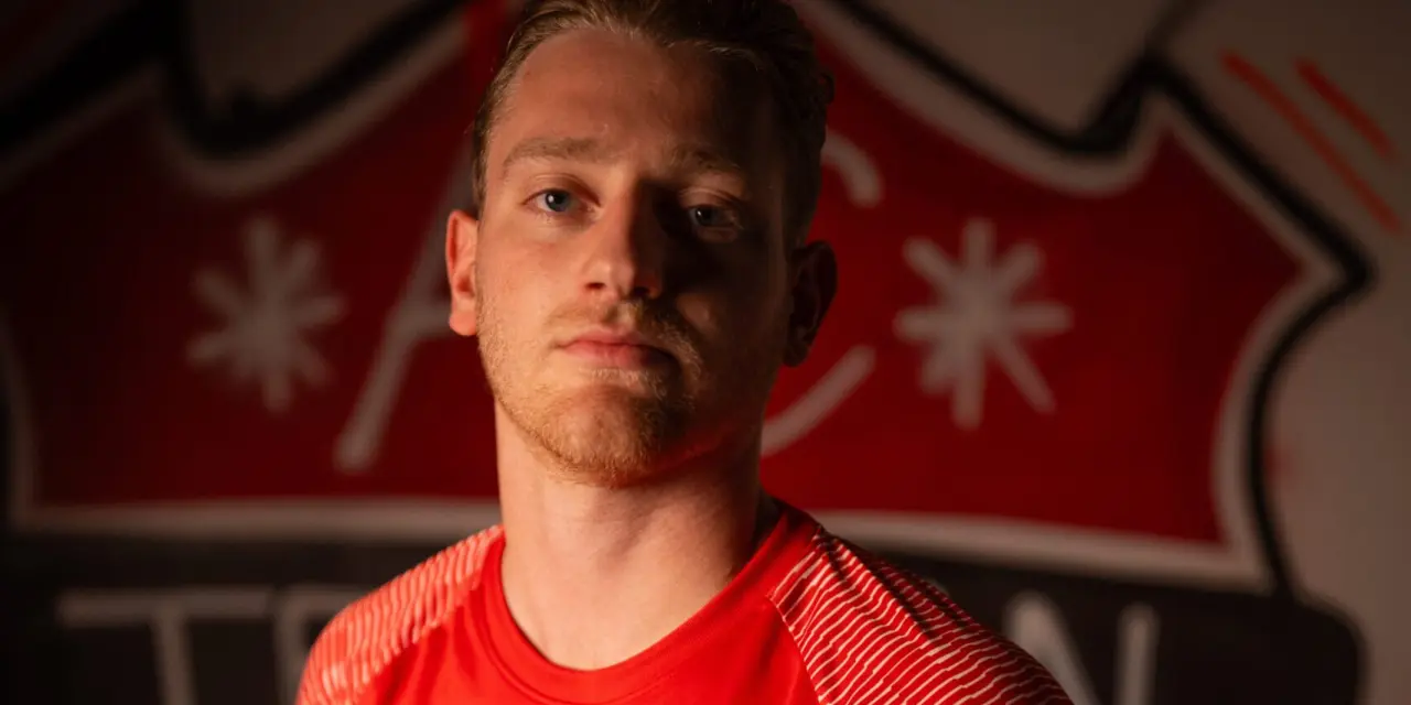 Spieler des AFC Terlan im roten Trikot mit Vereinswappen und Sponsorlogo vor einer großen Wappen-Wand, ernst in die Kamera blickend.