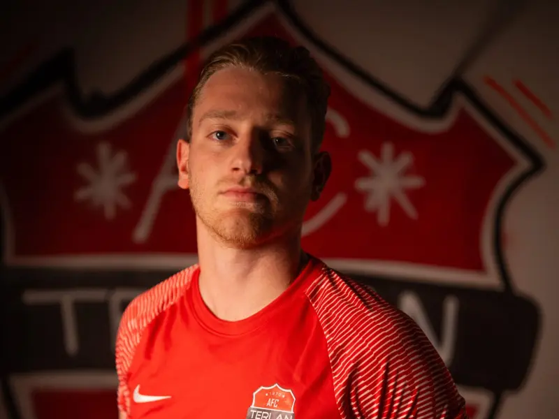 Spieler des AFC Terlan im roten Trikot mit Vereinswappen und Sponsorlogo vor einer großen Wappen-Wand, ernst in die Kamera blickend.