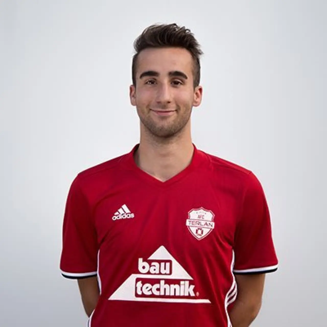 Porträt eines lächelnden jungen Mannes im roten Trikot des AFC Terlan mit Vereinswappen und bautechnik-Sponsorenlogo vor hellem Hintergrund.