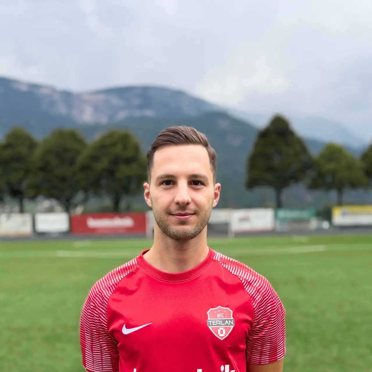 Porträt eines Spielers des AFC Terlan in roter Trikot mit Vereinswappen und bautechnik-Sponsor, vor einem Kunstrasenplatz und Bergen.