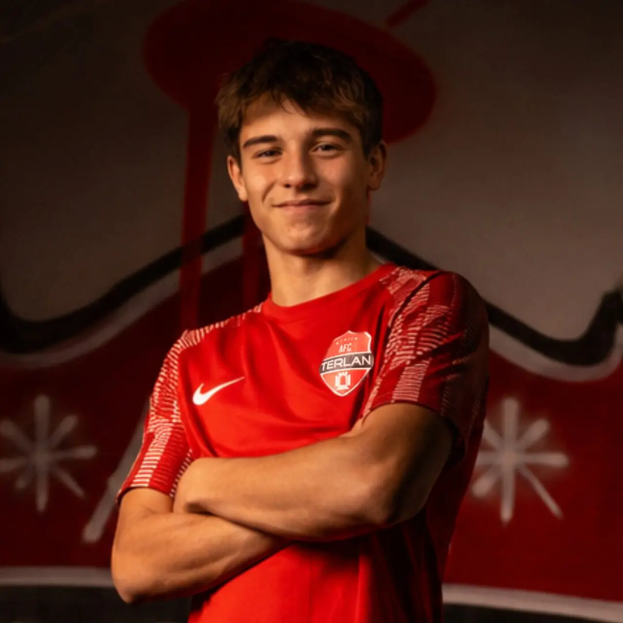 Junger Fußballer im roten Trikot des AFC Terlan mit verschränkten Armen lächelt vor einer roten Graffiti-Wand in die Kamera.