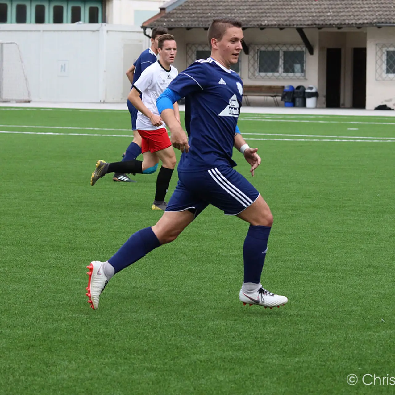 Spieler in dunkelblauem Trikot läuft auf dem Kunstrasen im Spiel, Gegenspieler in Weiß und Torhüter im Hintergrund.