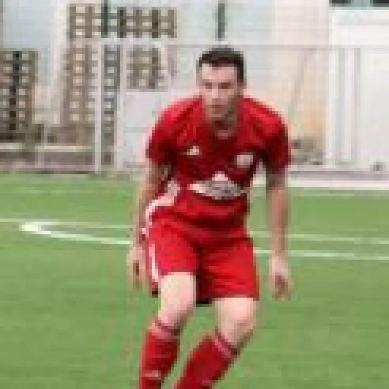 Ein Spieler in roter Vereinsschrift läuft auf einem grünen Kunstrasenplatz mit dem Ball am Fuß, im Hintergrund eine Hecke.