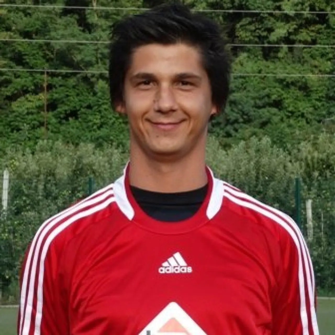 Porträt eines lächelnden jungen Mannes mit dunklen Haaren im roten Trikot mit Sponsorlogo bautechnik vor grünem Hintergrund.