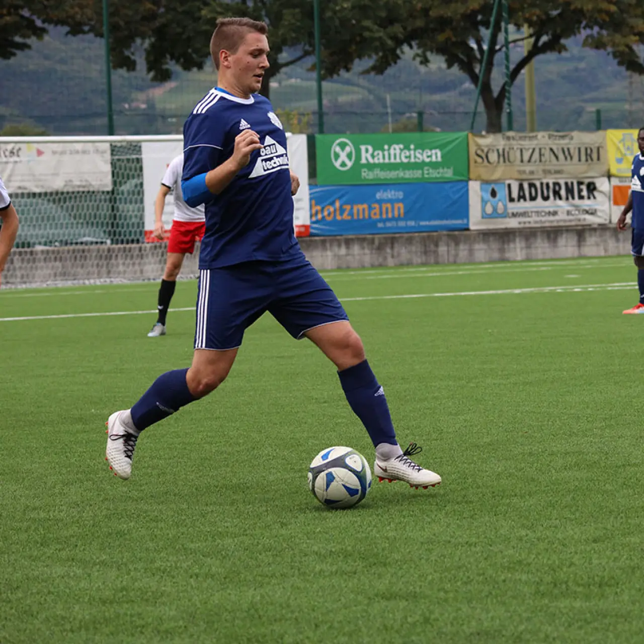 Spieler in blauem Trikot führt den Ball auf dem Kunstrasen, im Hintergrund Bandenwerbung von Raiffeisen, Holzmann und Zimmer.