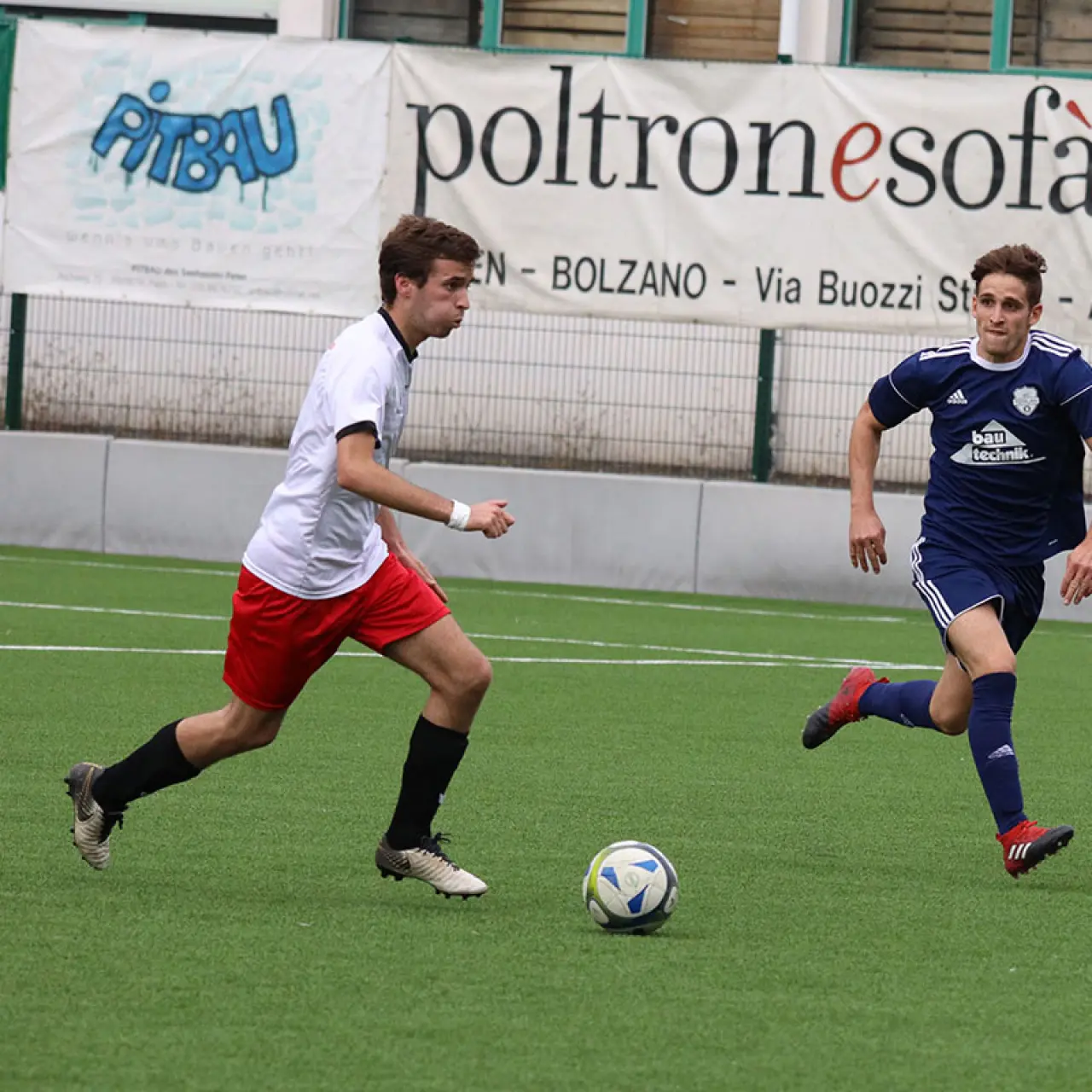 Zwei Spieler im Zweikampf um den Ball auf dem Kunstrasen: links in Weiß mit roter Hose, rechts in Dunkelblau des AFC Terlan.