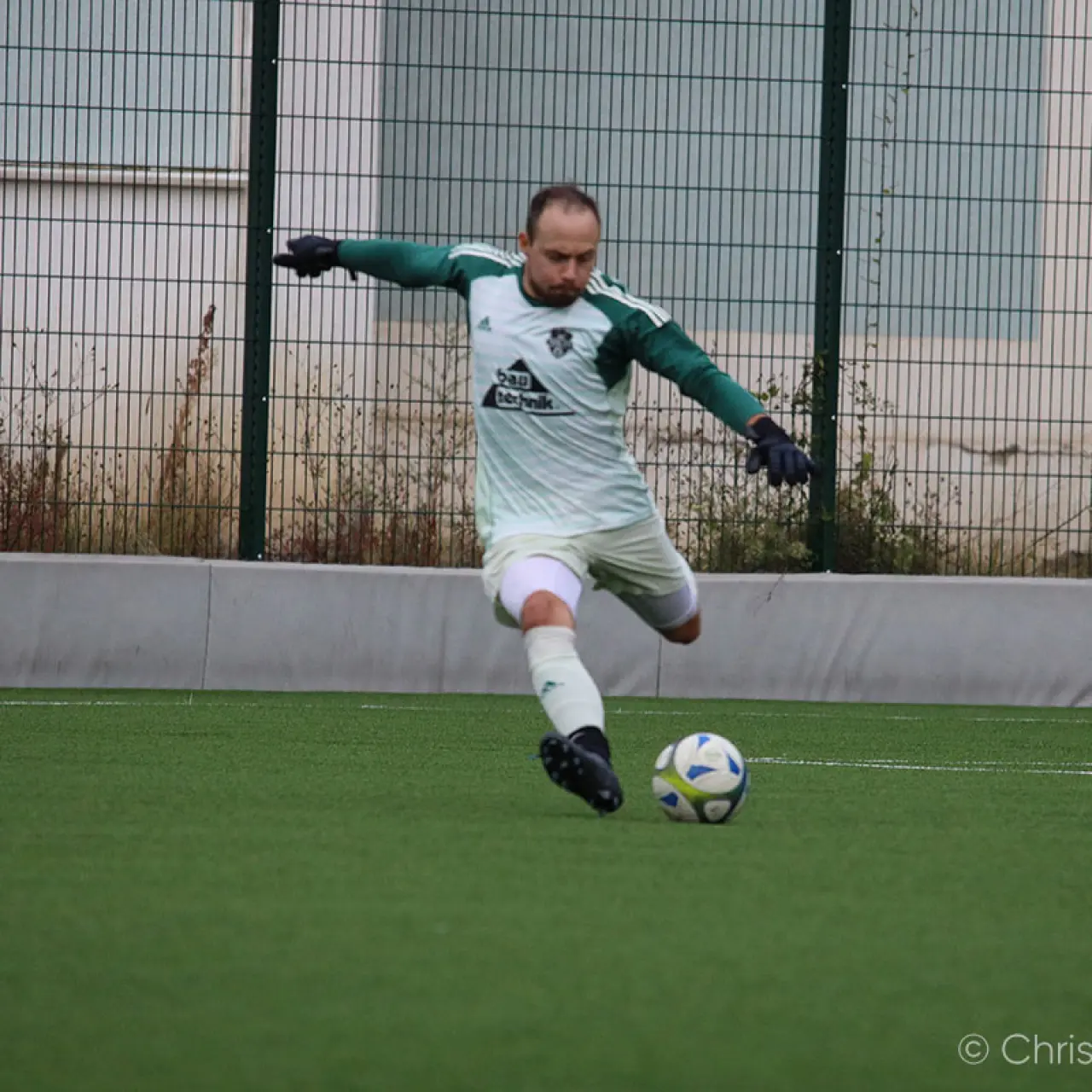 Ein Spieler in weiß-grünem Trikot mit grünen Handschuhen schießt auf dem Kunstrasenplatz einen Ball, im Hintergrund ein Metallzaun.