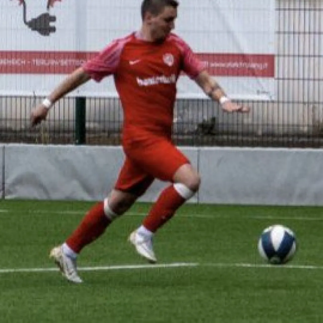 Spieler in roter Trikotkombination führt den Ball auf dem Kunstrasen, im Hintergrund Werbebande von Elektro Lang.
