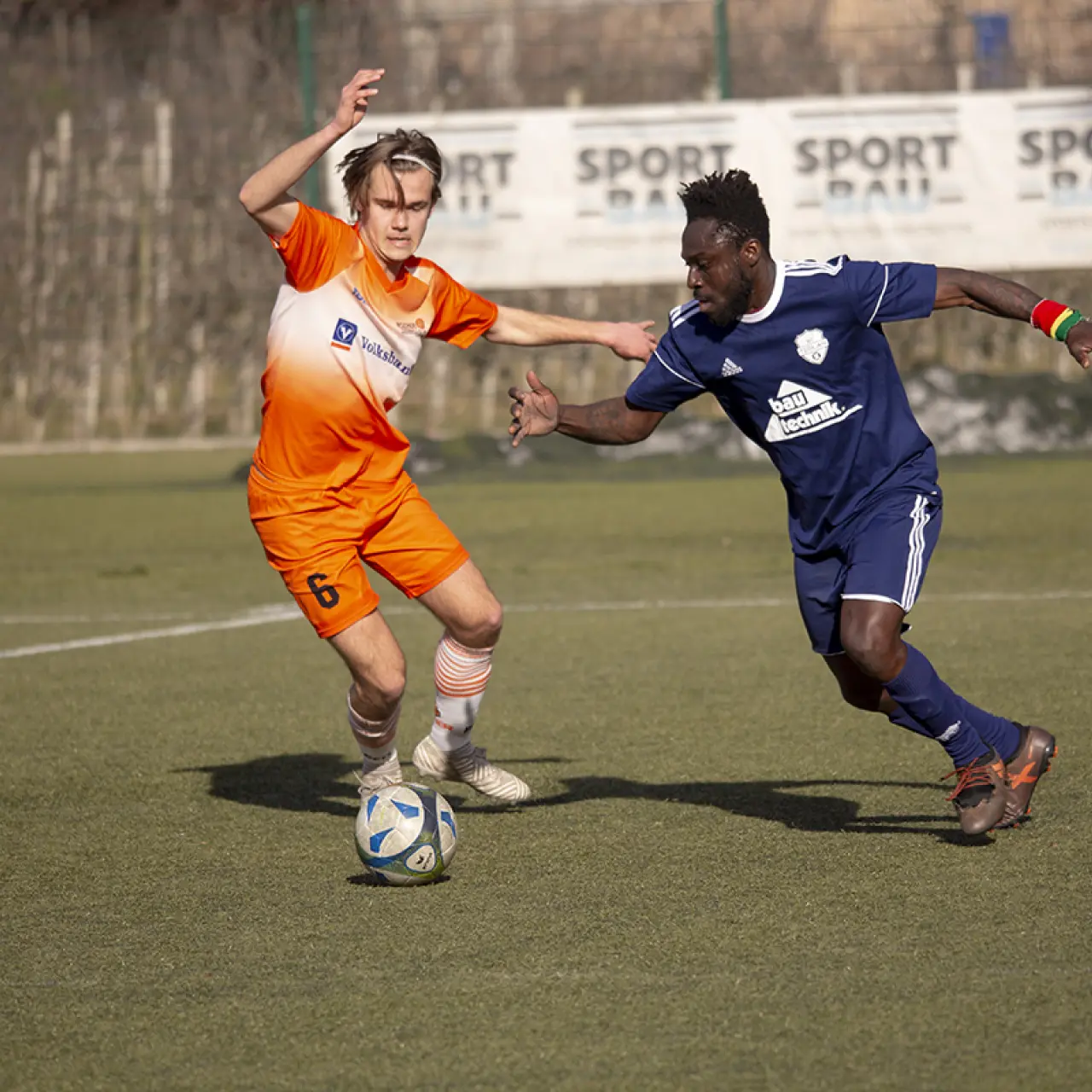 Zweikampf auf dem Rasen: Ein Spieler in Orange mit der Nummer 6 führt den Ball, ein Gegenspieler in Dunkelblau versucht ihn zu stoppen.