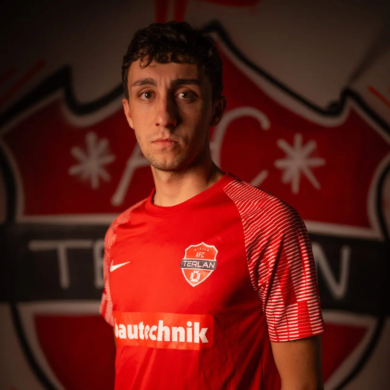 Spieler des AFC Terlan im roten Trikot mit Vereinswappen und Bautechnik-Sponsor, vor einer roten Wand mit dem Vereinslogo.