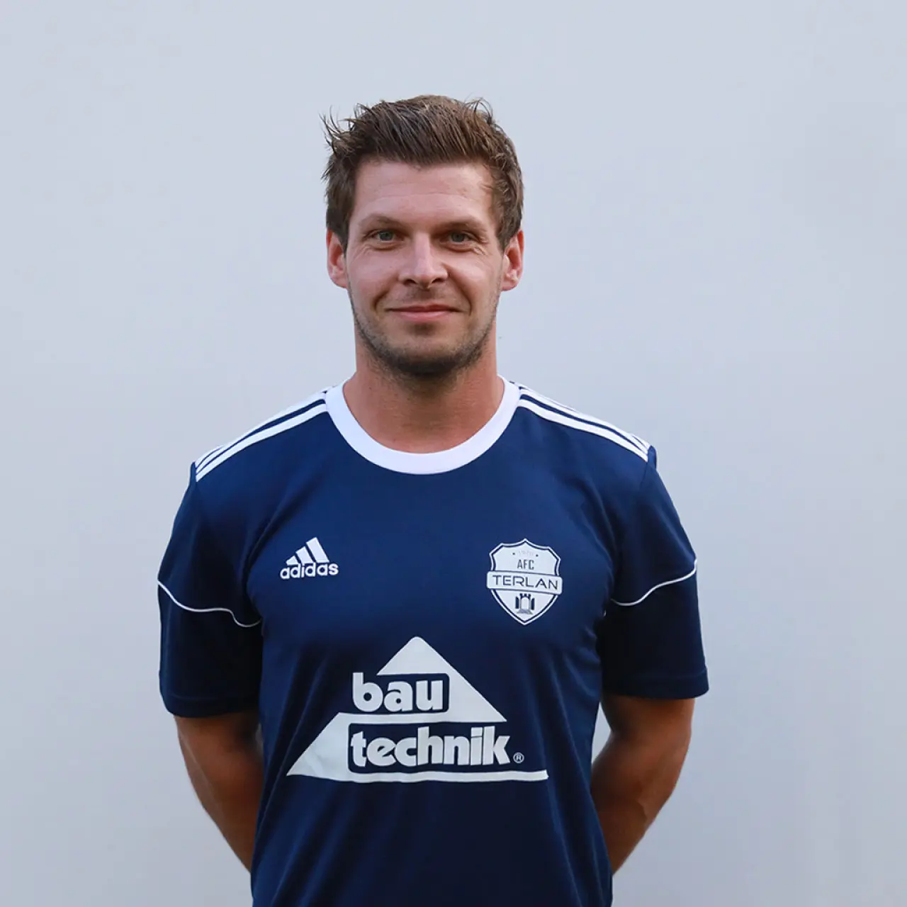 Porträtfoto eines lächelnden Mannes mit kurzen braunen Haaren in dunkelblauem AFC-Terlan-Trikot mit bautechnik-Sponsorlogo vor grauem Hintergrund.