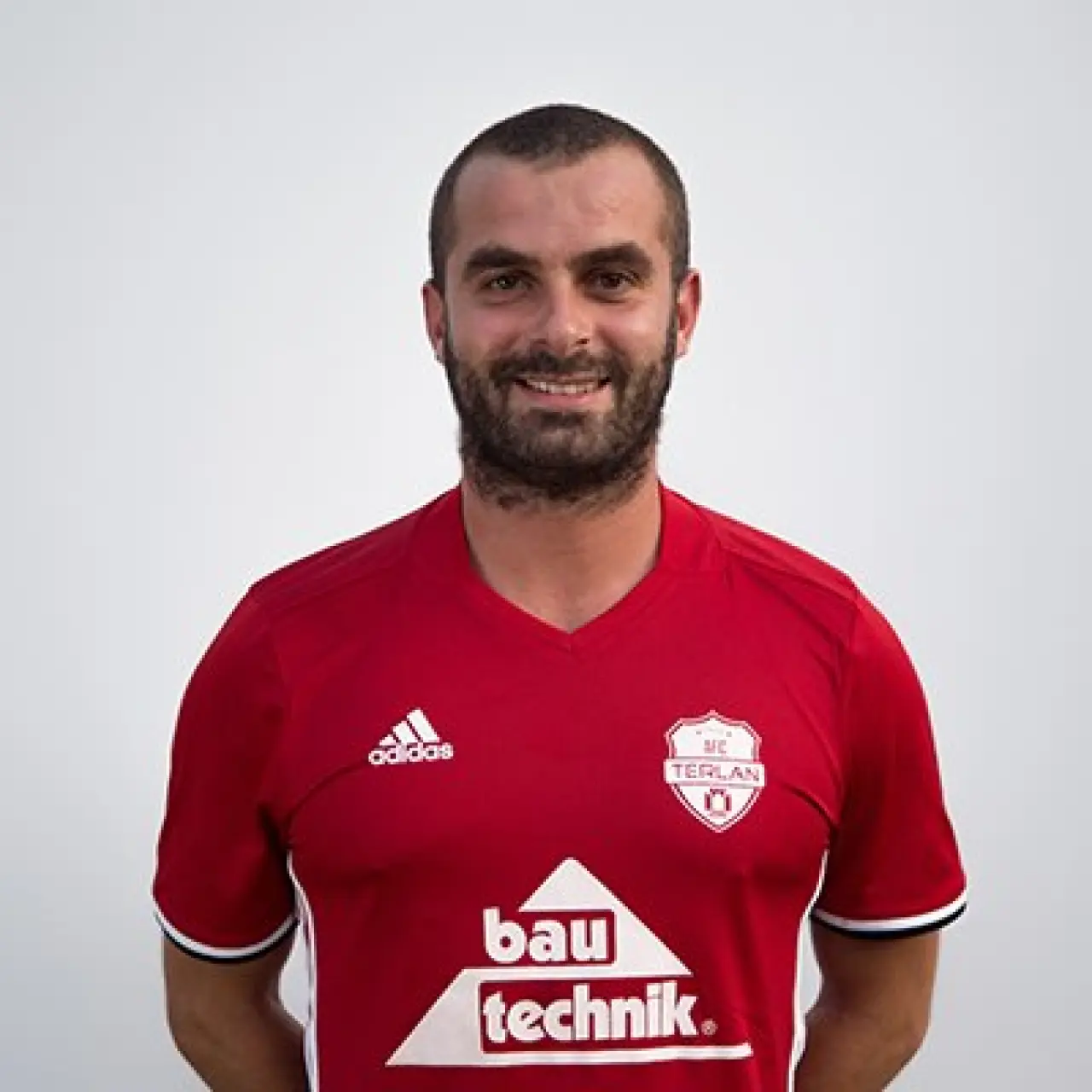 Porträt eines lächelnden Fußballspielers mit Bart im roten Trikot des AFC Terlan mit bautechnik-Sponsorenlogo vor hellgrauem Hintergrund.