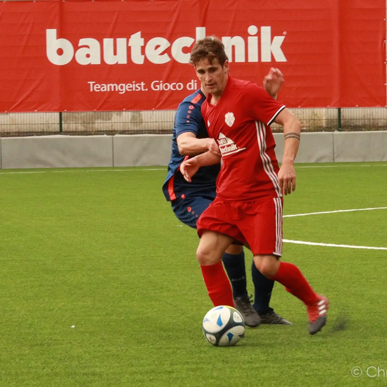 Ein Spieler in roter Trikot mit weißen Streifen führt den Ball auf dem Kunstrasen, ein Gegenspieler in Dunkelblau folgt, im Hintergrund Werbebanner von bautechnik.
