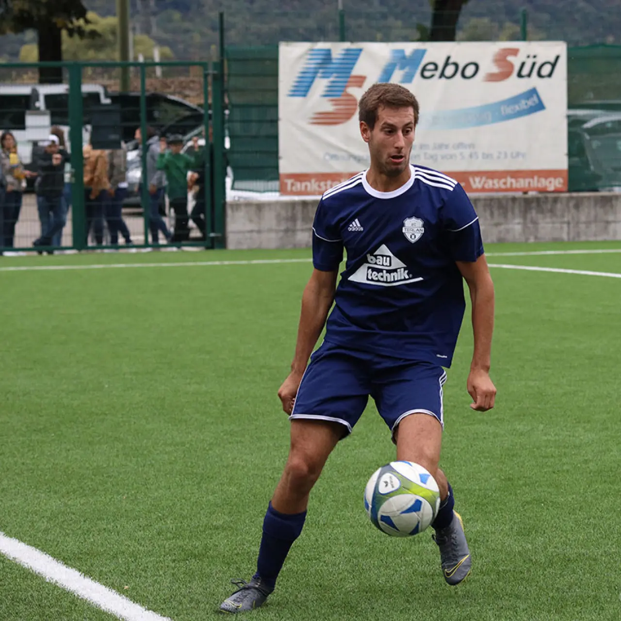 Spieler in blauem Trikot mit Vereinswappen führt den Ball auf dem Kunstrasen, im Hintergrund Banner von Mebo Süd und Zuschauer am Zaun.