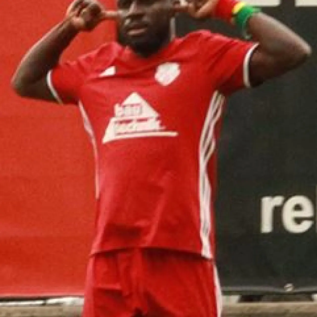 Fußballspieler in roten Trikot und Stutzen jubelt auf dem Rasen, beide Hände neben dem Kopf, am Arm eine bunte Binde.