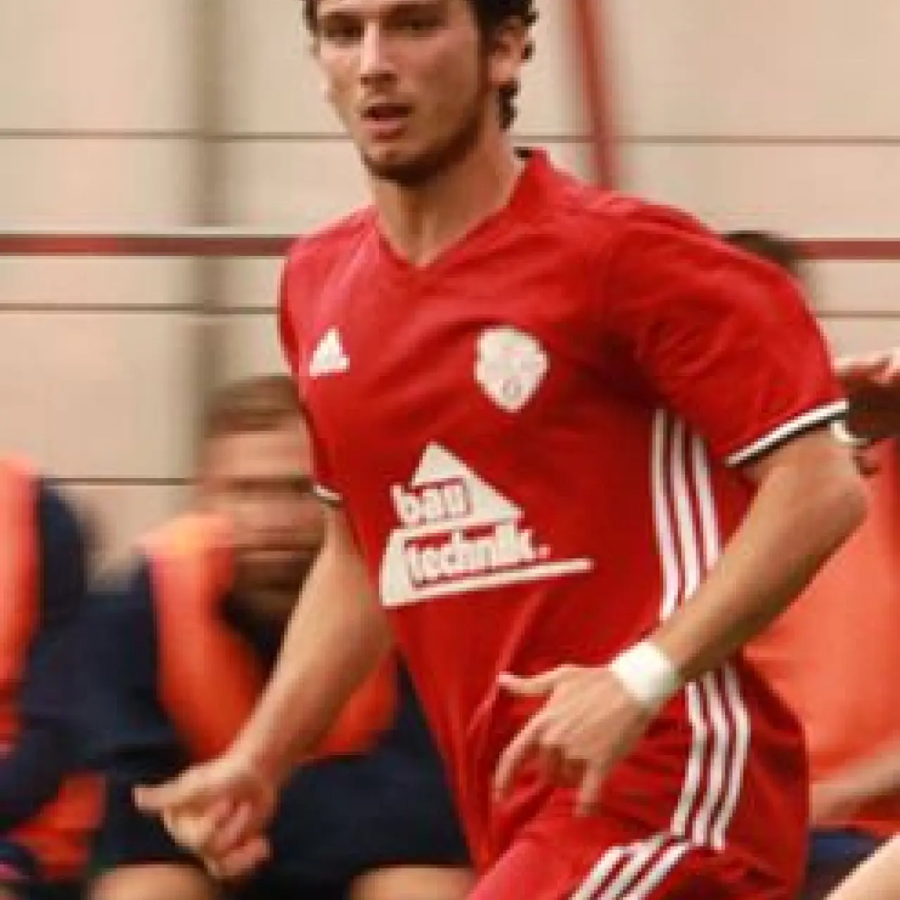 Spieler Alession Perri in roter Trikot-und-Stutzen-Kombi mit Bautechnik-Sponsor läuft am Ball über den Rasen bei einer Spielszene des AFC Terlan.