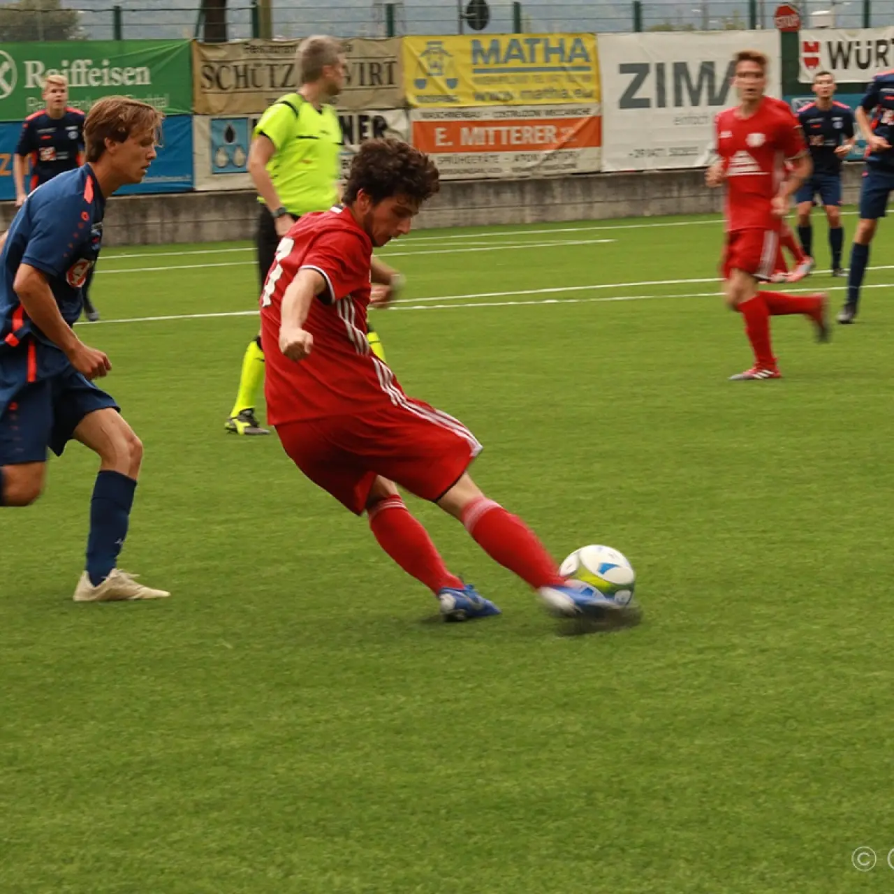 Spielszene: Ein Spieler in Rot schießt den Ball auf dem Rasen, während ein Gegenspieler in Blau und Banden mit Sponsorennamen im Hintergrund zu sehen sind.