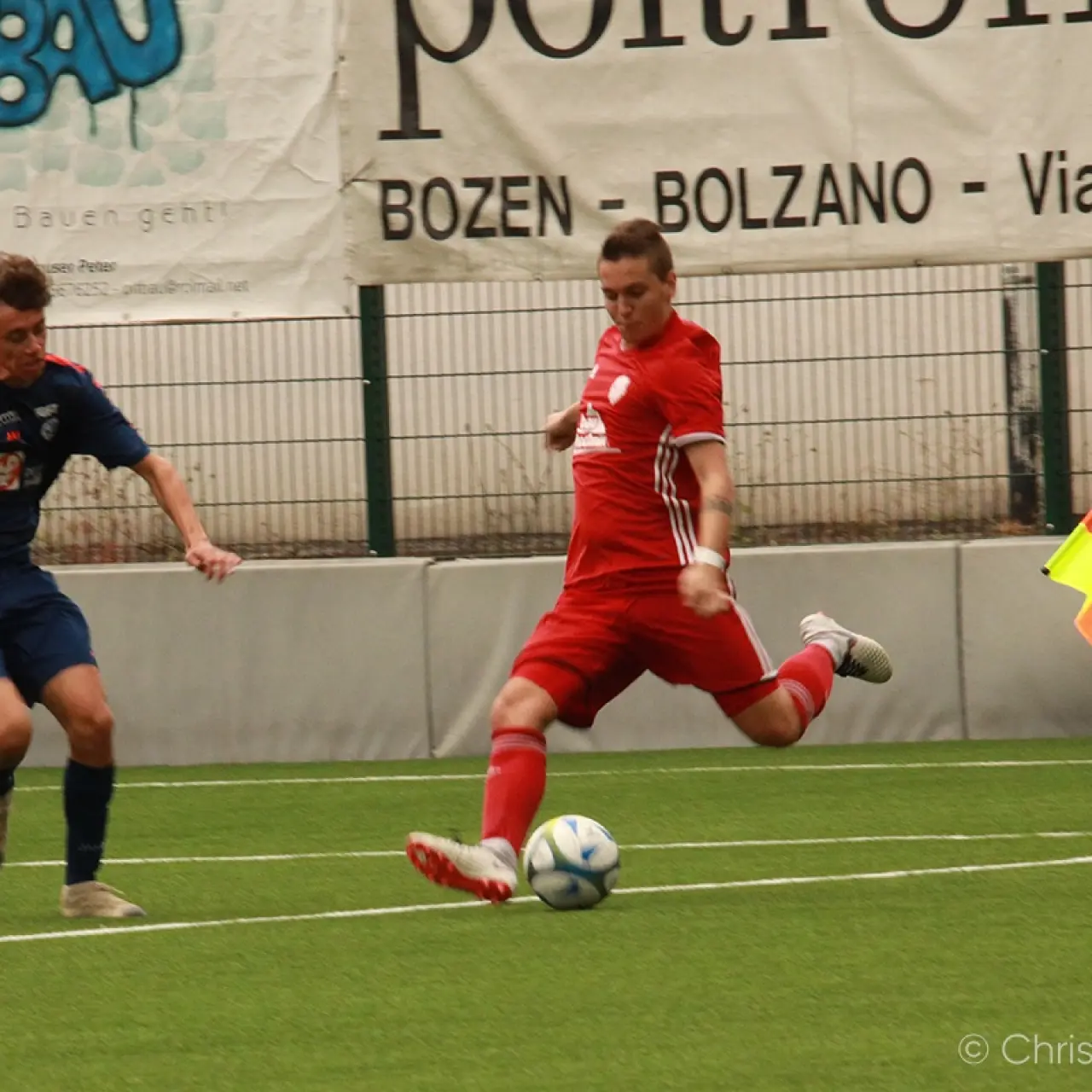 Spielszene auf Kunstrasen: Ein Spieler in Rot holt zum Schuss aus, ein Gegner in Dunkelblau setzt nach, im Hintergrund ein Schiedsrichterassistent mit Fahne.