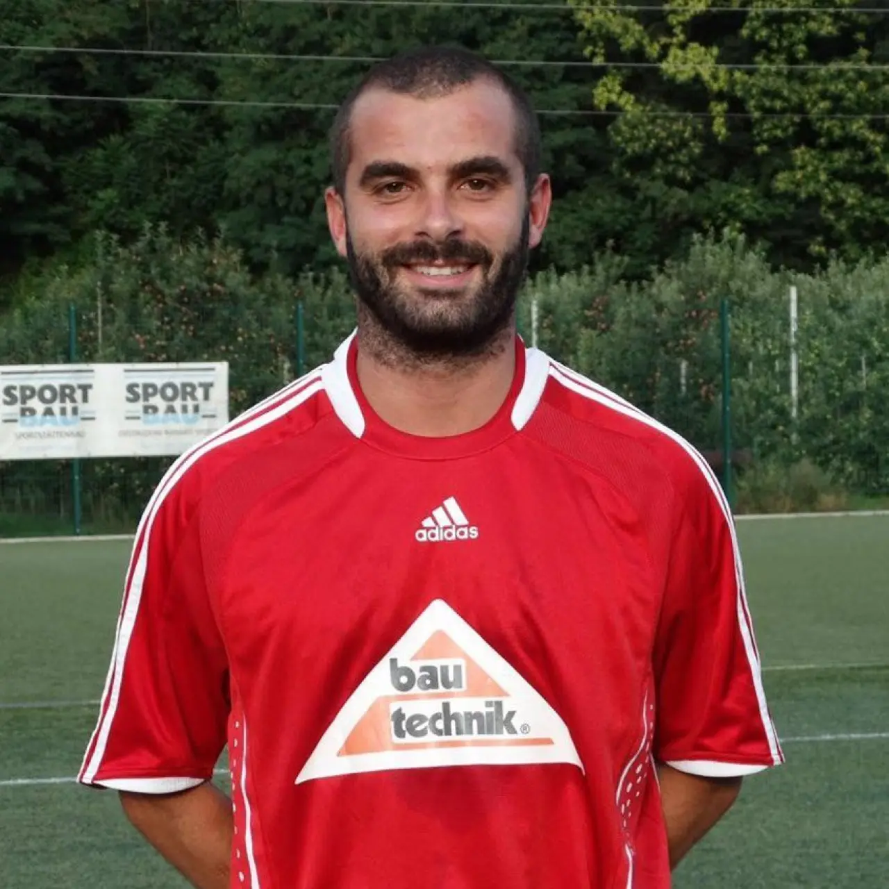Porträt eines lächelnden Fußballspielers mit Bart im roten Trikot mit bautechnik-Sponsorlogo vor einem Kunstrasenplatz.