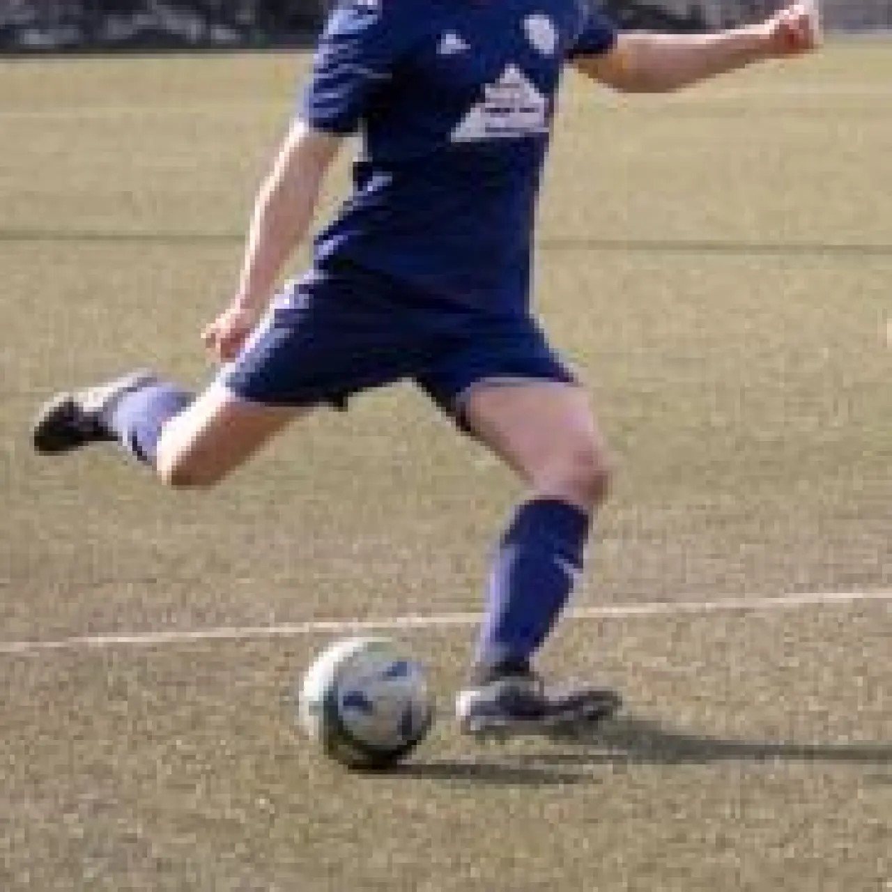 Ein Spieler in blauem Trikot holt auf dem Kunstrasen zum Schuss aus, der Ball liegt vor ihm am Boden, im Hintergrund Werbebanden.
