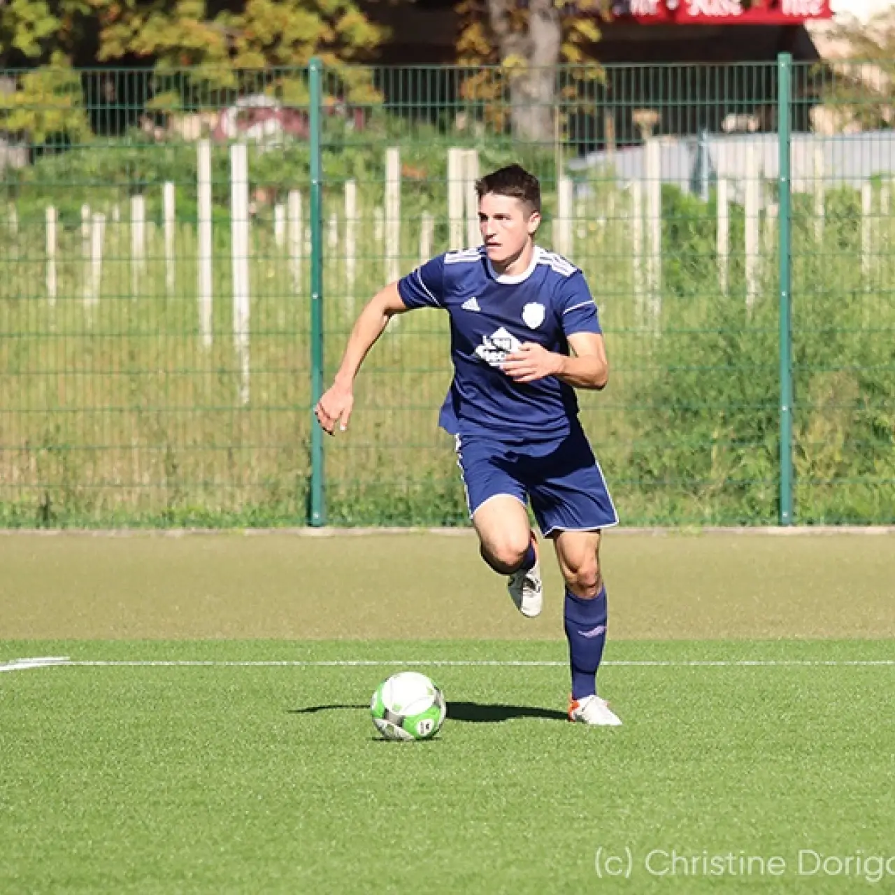 Spieler in dunkelblauem Trikot führt den Ball auf dem Kunstrasen, im Hintergrund ein Gegenspieler in Rot-Weiß.