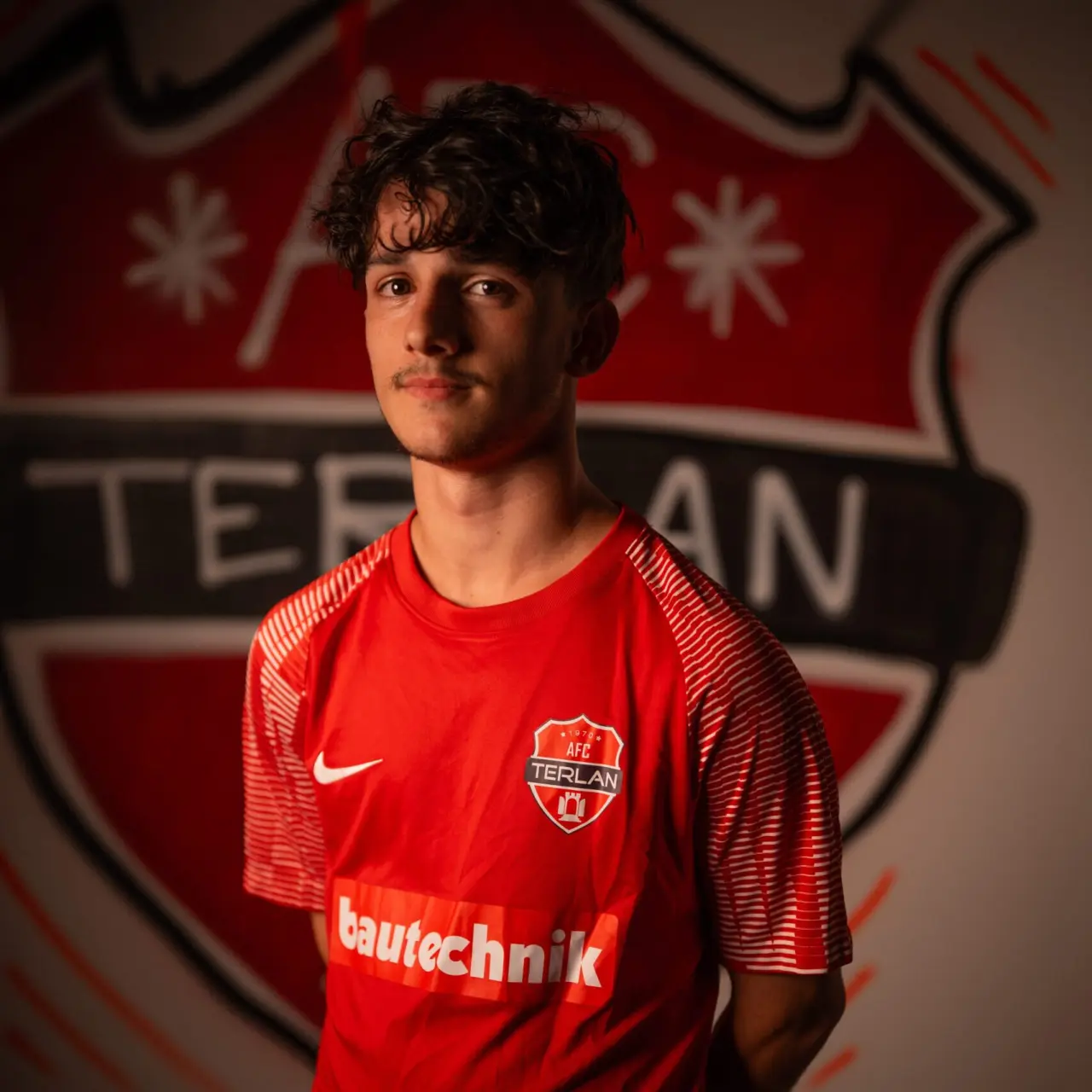 Junger Spieler im roten AFC-Terlan-Trikot mit Bautechnik-Sponsor vor dem bemalten Vereinswappen des AFC Terlan.