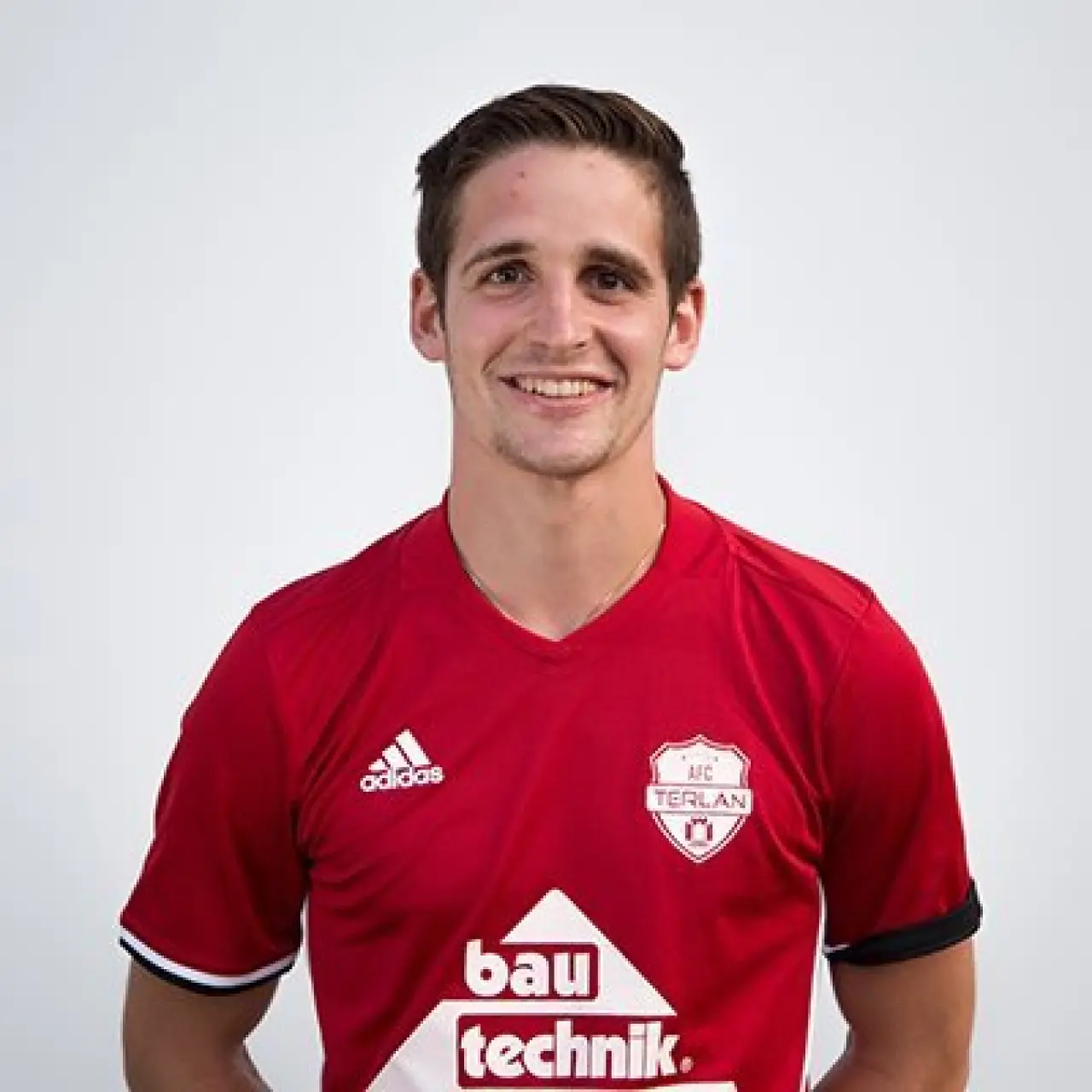 Porträtfoto von Elias Erschbamer im roten Trikot des AFC Terlan mit Vereinswappen, vor hellem Hintergrund, lächelnd.