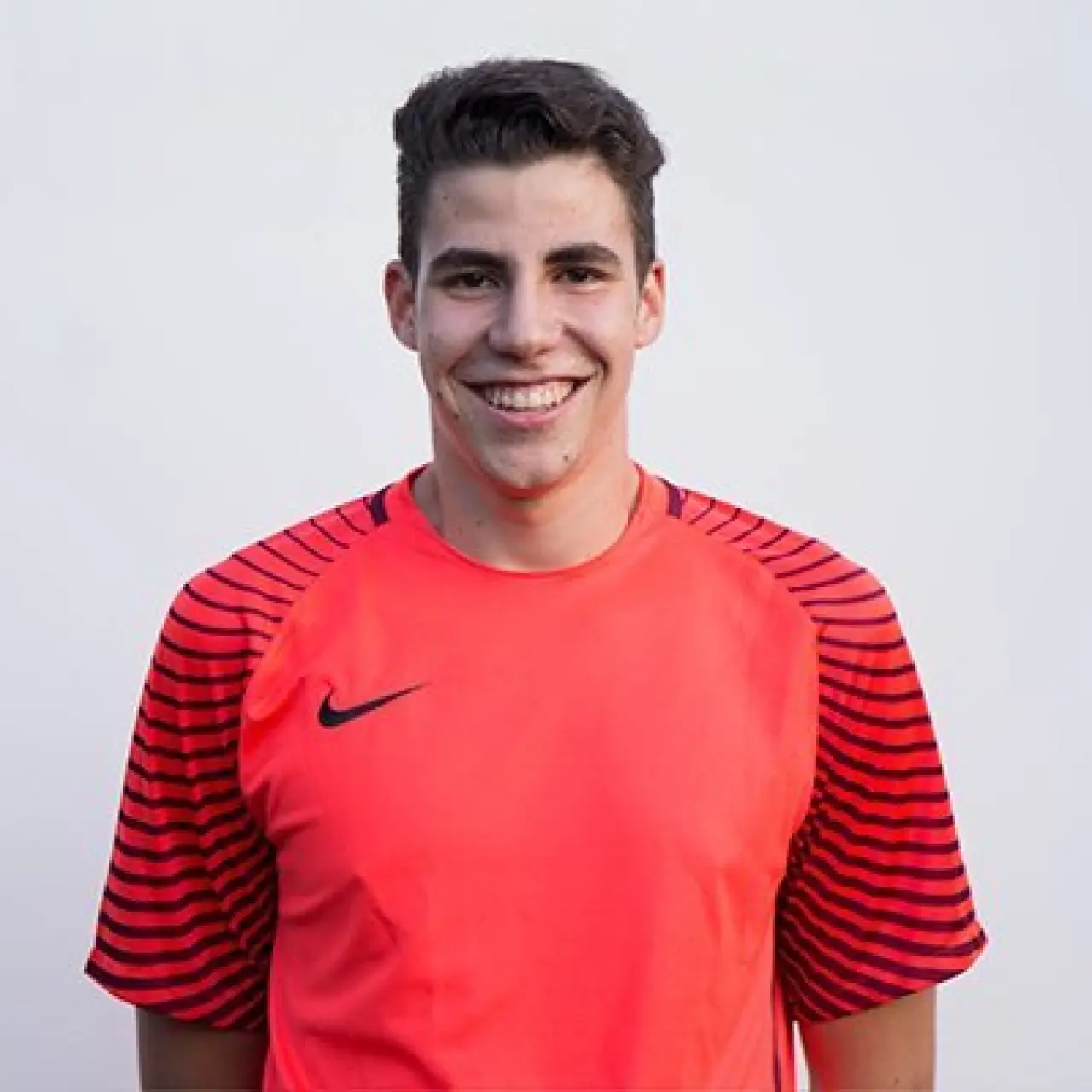 Porträt eines lächelnden jungen Fußballspielers in orangeroter Trikot mit Nike-Logo vor hellem Hintergrund.