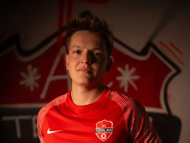 Porträt eines Spielers des AFC Terlan im roten Trikot mit Vereinswappen und Bautechnik-Sponsor vor einer Wand mit Vereinslogo.