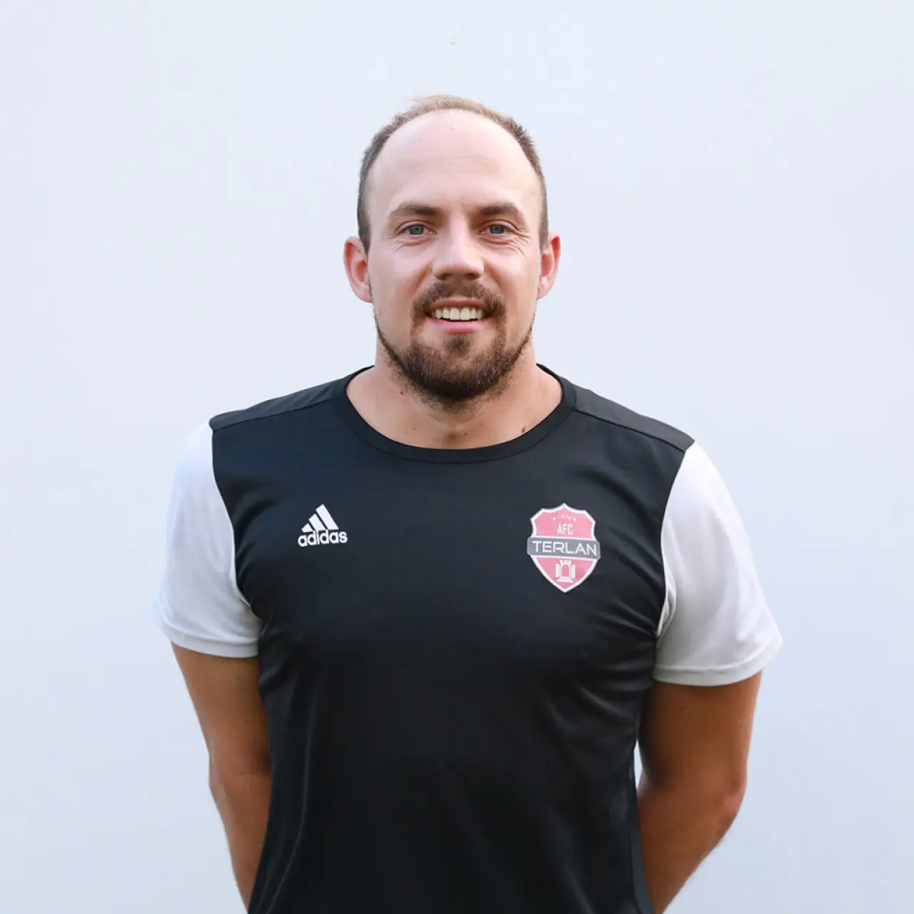 Porträtfoto eines lächelnden Mannes mit Bart in schwarzem Trikot mit AFC Terlan Wappen vor weißer Wand.