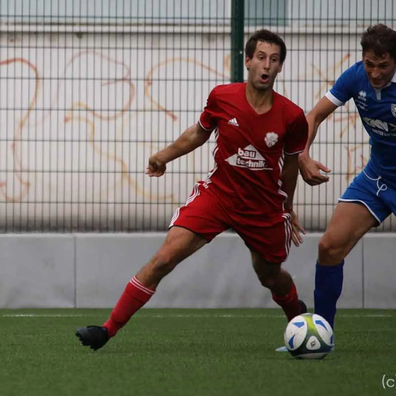 Spielszene: Ein Spieler in Rot von Bau-Technik verfolgt den Ball gegen einen Gegenspieler in Blau auf dem Kunstrasenplatz, AFC Terlan gegen Bruneck.