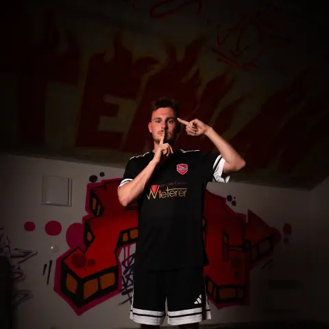 Andreas Walcher in schwarzem AFC-Terlan-Trikot vor roter Graffiti-Wand, Finger an den Lippen und an der Schläfe.