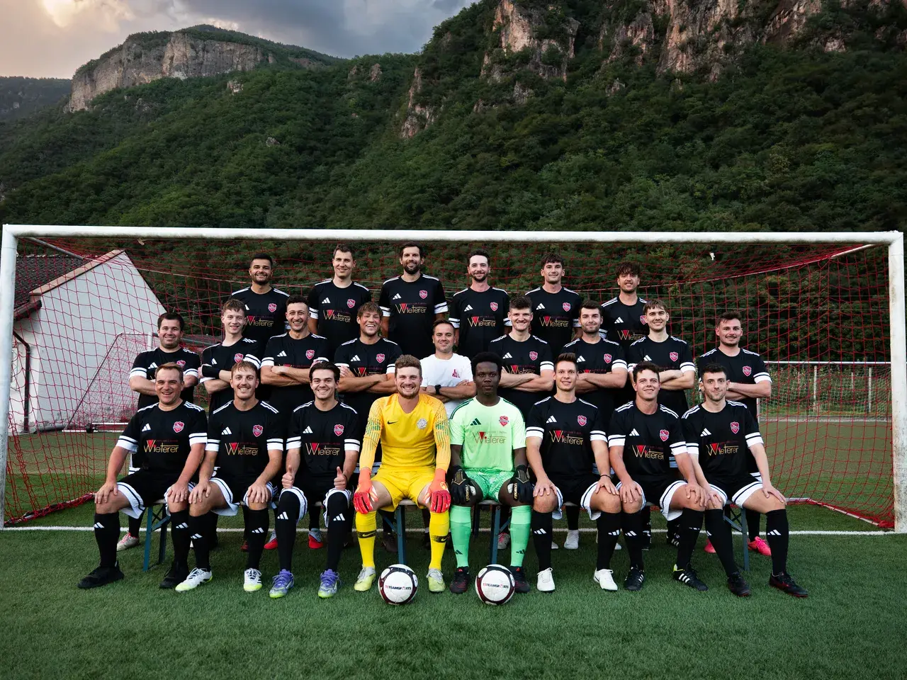 Mannschaftsfoto der Sturmtruppen des AFC Terlan in schwarzen Trikots mit Sponsorenaufdruck vor einem Tor, davor Berge.