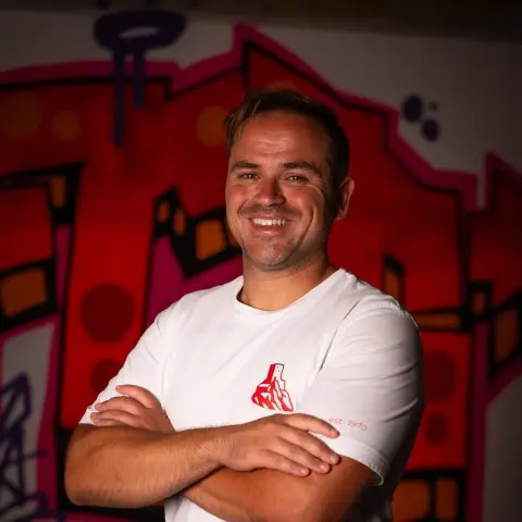 Lächelnder Mann mit verschränkten Armen vor einer roten Graffiti-Wand, im weißen Shirt mit rotem AFC Terlan-Logo und Schriftzug "est. 1970".