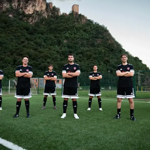 Sieben Spieler des AFC Terlan in schwarzen Trikots mit weißen Streifen stehen mit verschränkten Armen auf dem Kunstrasen vor einer bewaldeten Felswand.